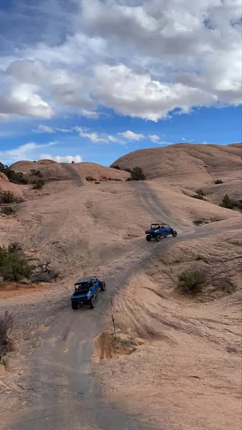 Two UTVs climbing a winding slickrock trail under a partly cloudy sky