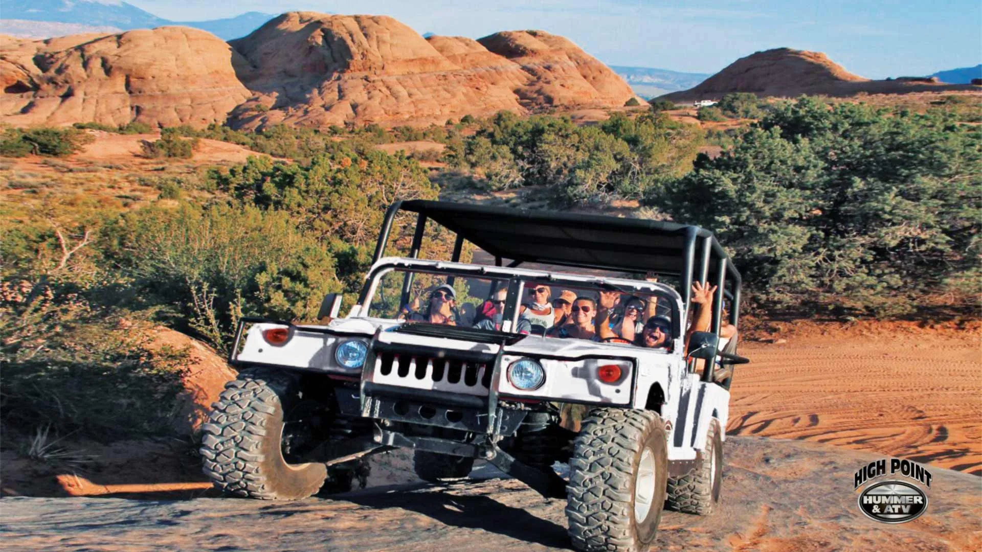 White open-top Hummer full of guests driving across red sand among junipers and sandstone domes