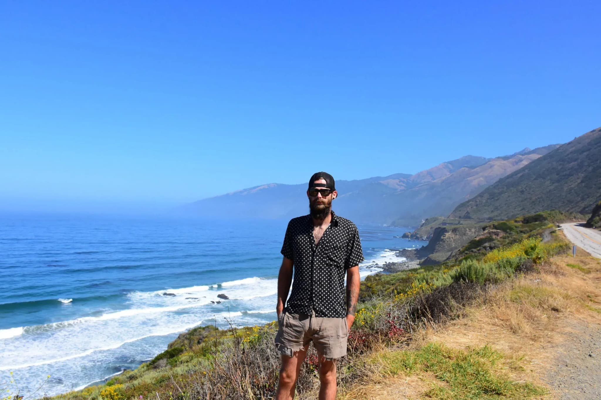 Man in sunglasses and a patterned shirt standing on a bluff above a rugged ocean coastline