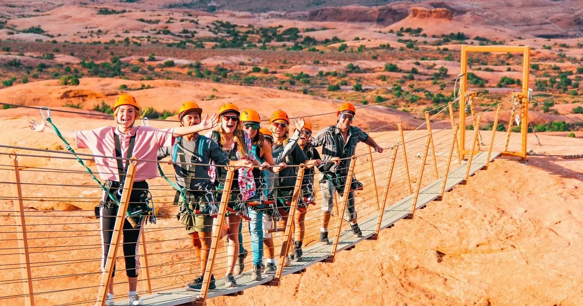 Guests in orange helmets crossing a suspension bridge above red-rock desert on the Raven's Rim zipline course