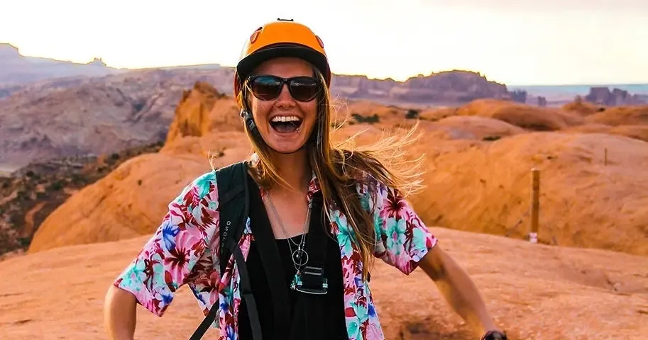 Guest in an orange helmet and floral shirt laughing on the red-rock zipline course at sunset