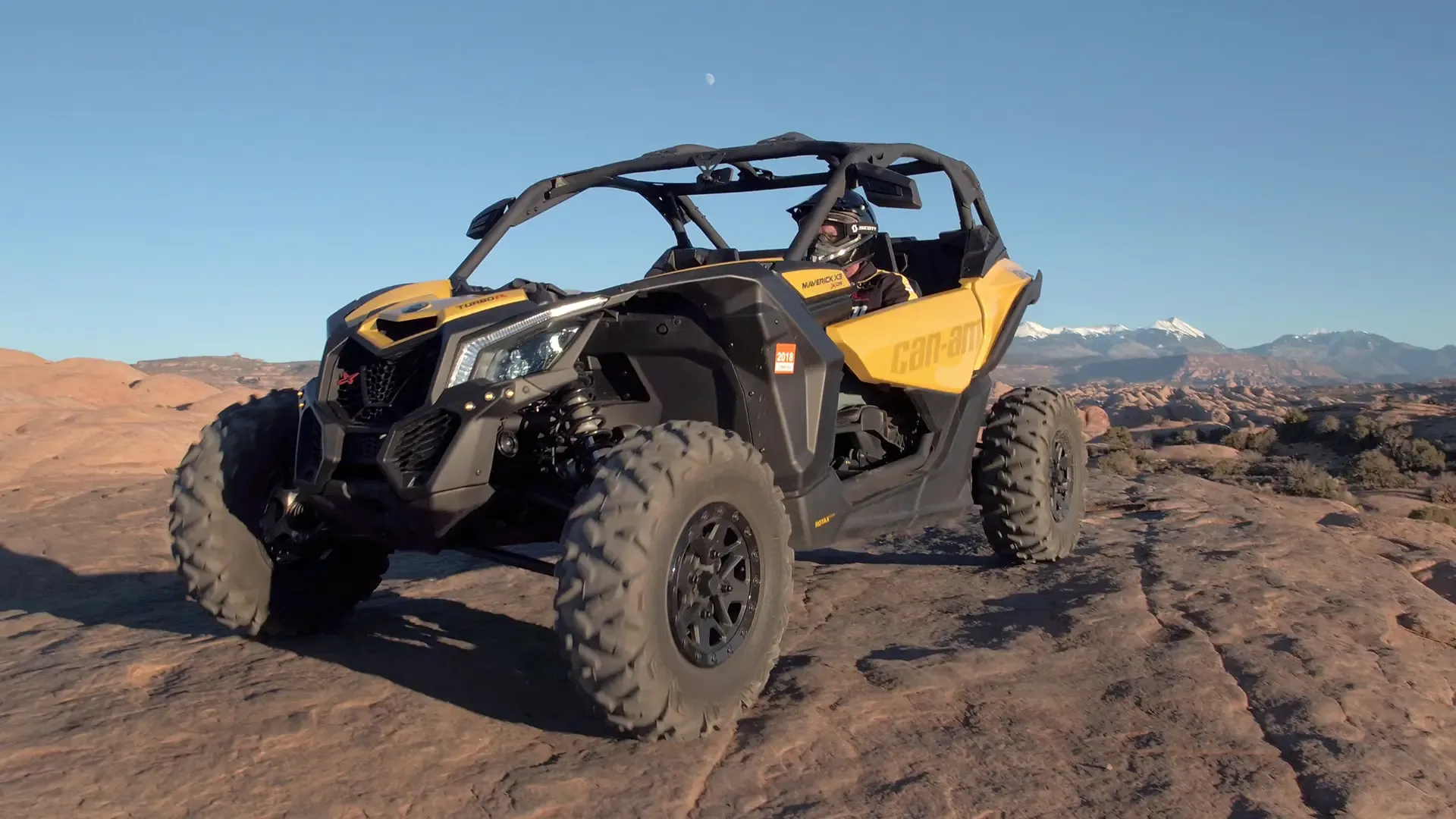 A bright yellow off-road vehicle sits on a rock surface at sunset, with distant snow-capped mountains.
