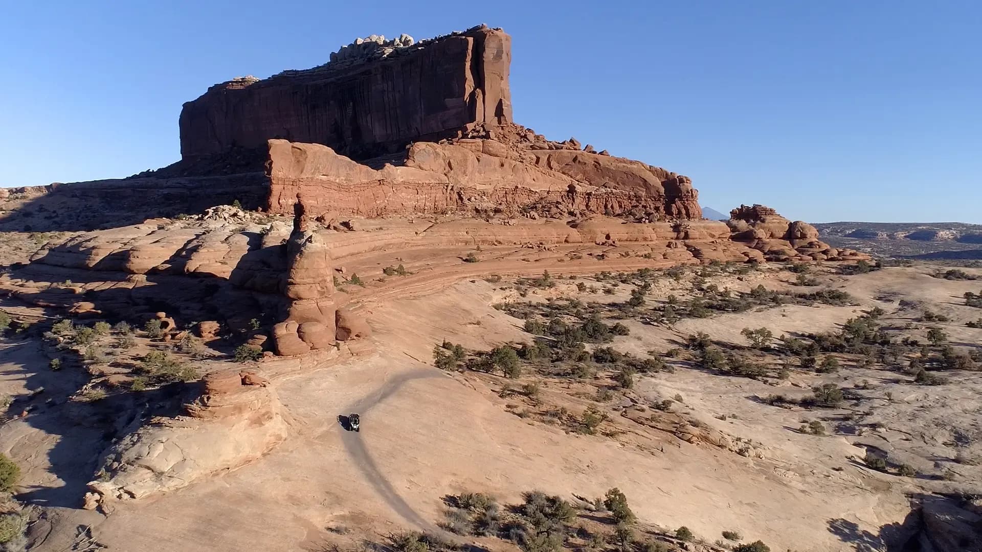 Aerial view of a UTV driving a desert trail beneath a large red rock butte near Monitor & Merrimac