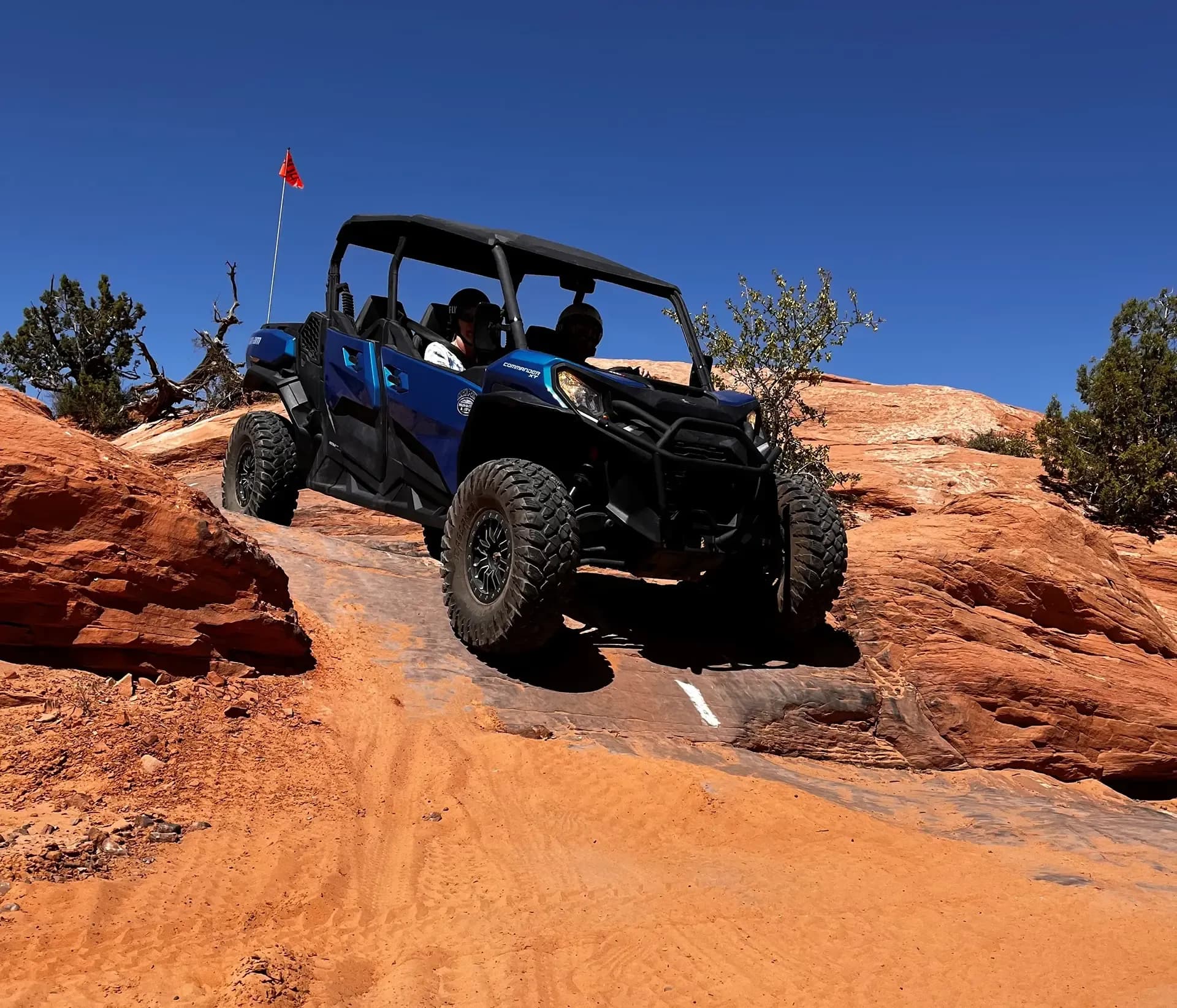 Blue Can-Am side-by-side easing down a red rock ledge with a red safety flag overhead