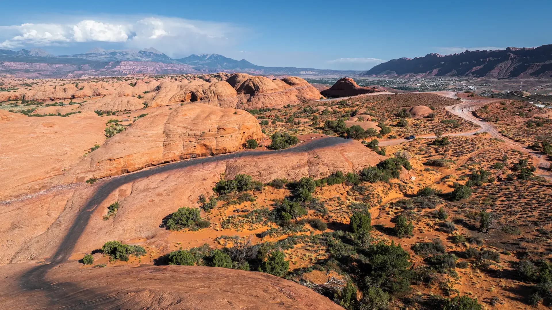 Aerial view of a dirt UTV trail winding through red slickrock domes above Moab, mountains in the distance