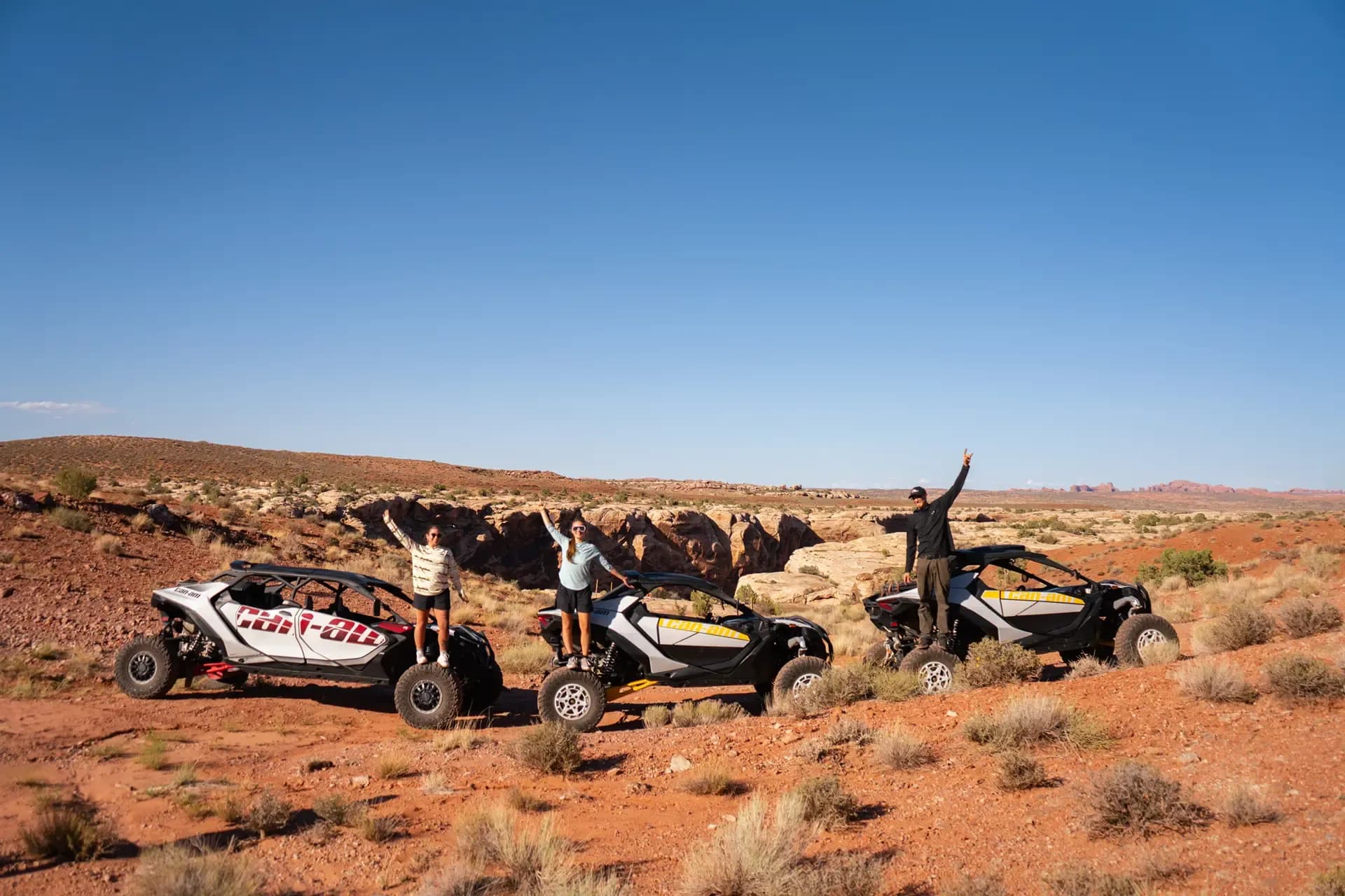 Riders celebrating beside three Can-Am side-by-sides parked on red desert sand