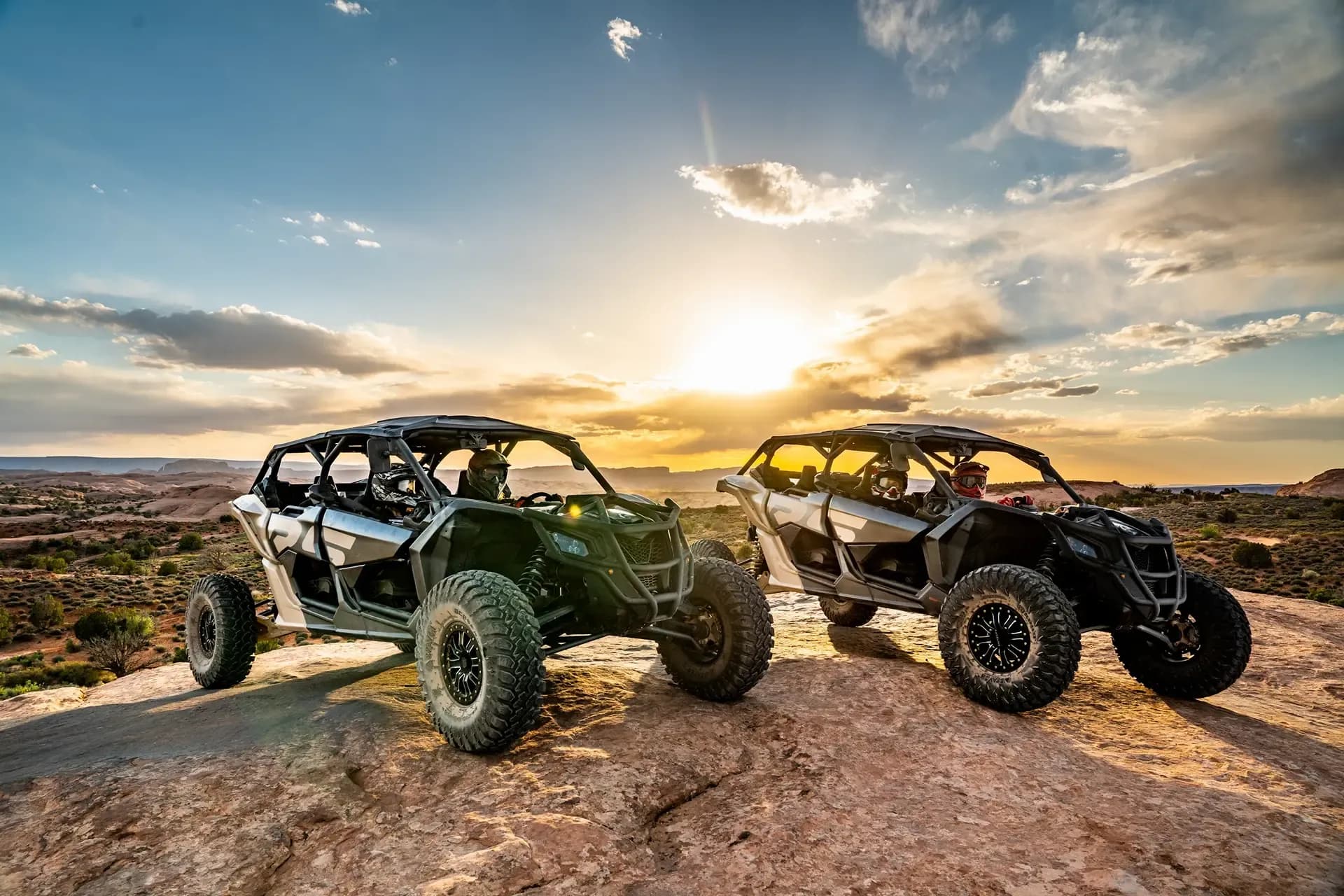 Two off-road vehicles parked on a rock outcrop at sunset, silhouetted against a brilliant sky.