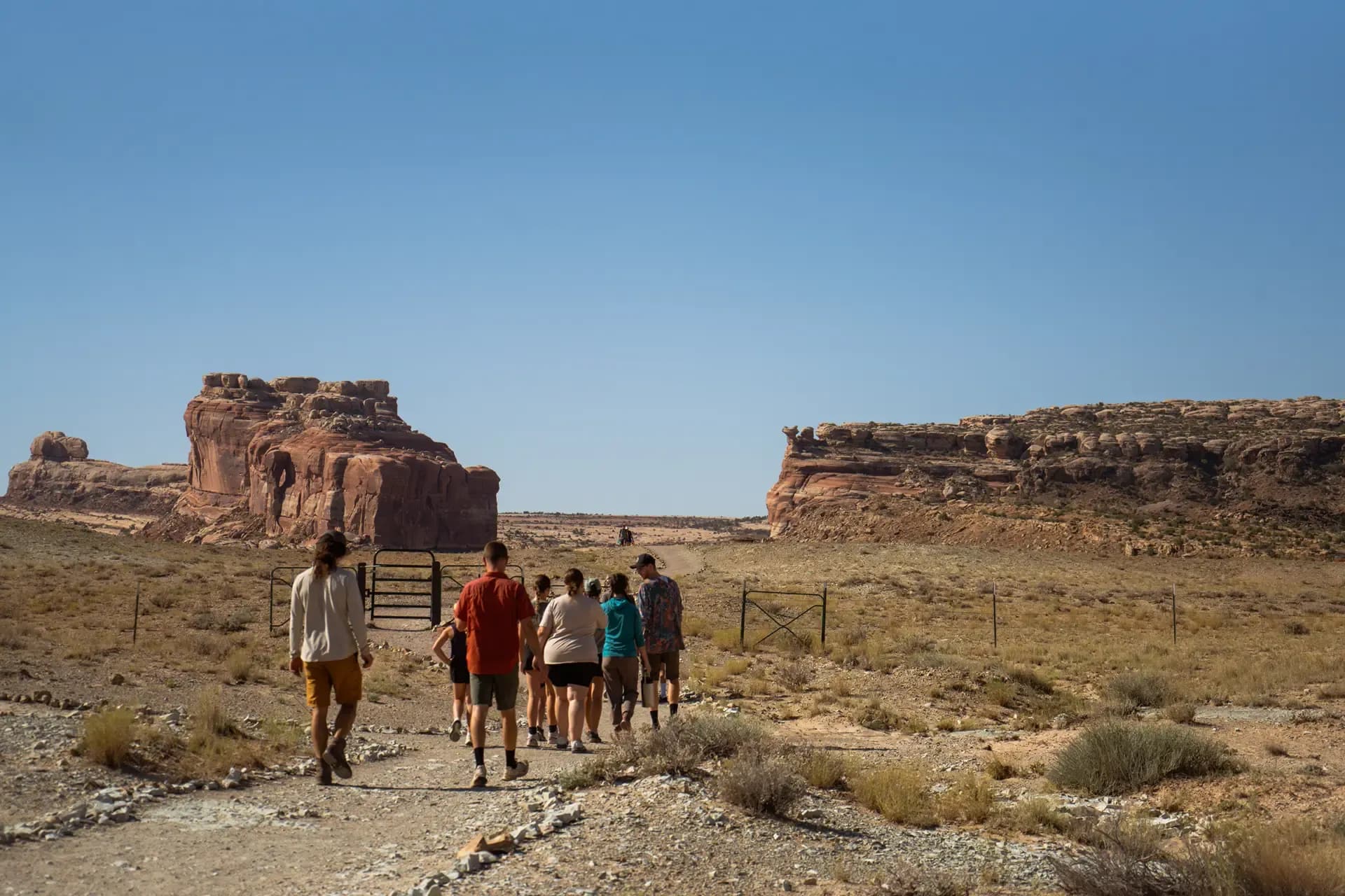 Hiking group walking a desert trail between red rock formations toward a trailhead gate