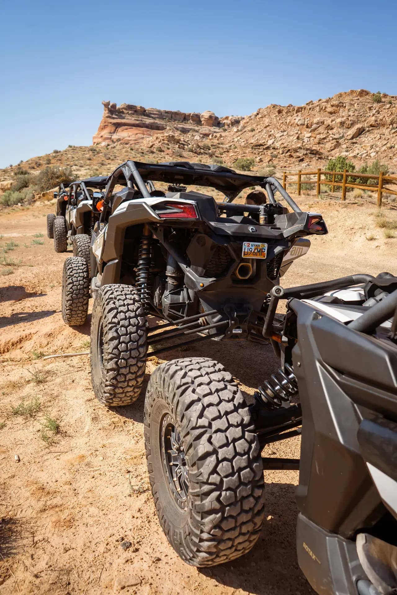 Rear of a Can-Am Maverick X3 with knobby tires parked on a dirt lot near red cliffs