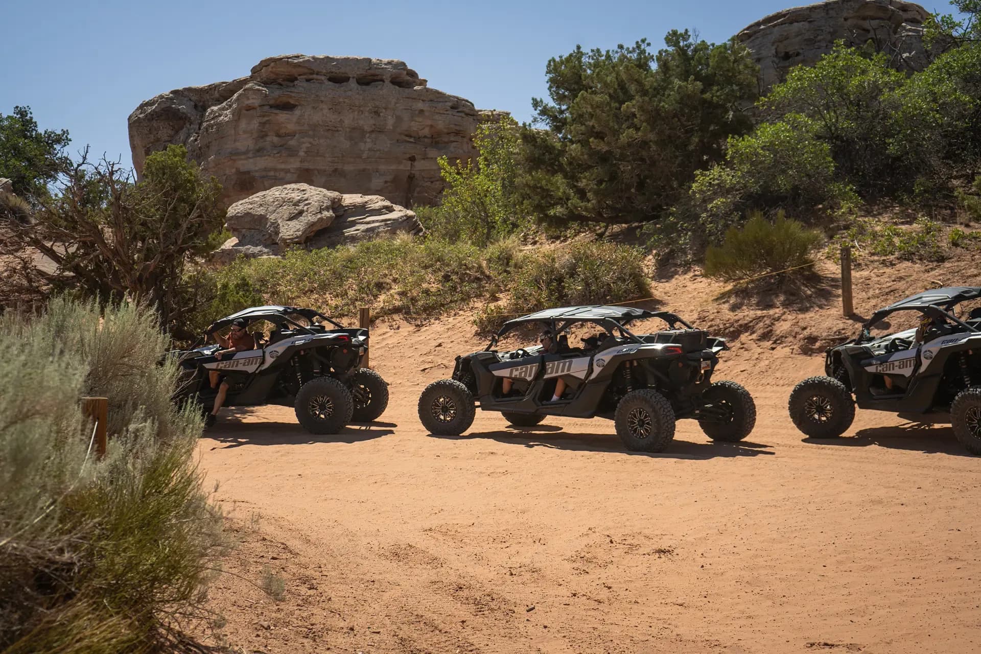 Three Can-Am side-by-sides parked in a row on red sand beside juniper and sagebrush
