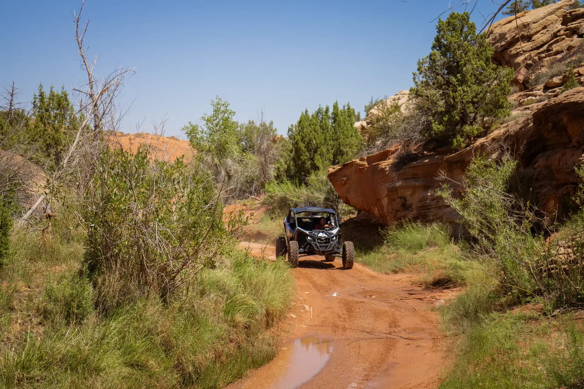 Can-Am Maverick X3 driving a sandy trail through green brush toward a red sandstone cliff