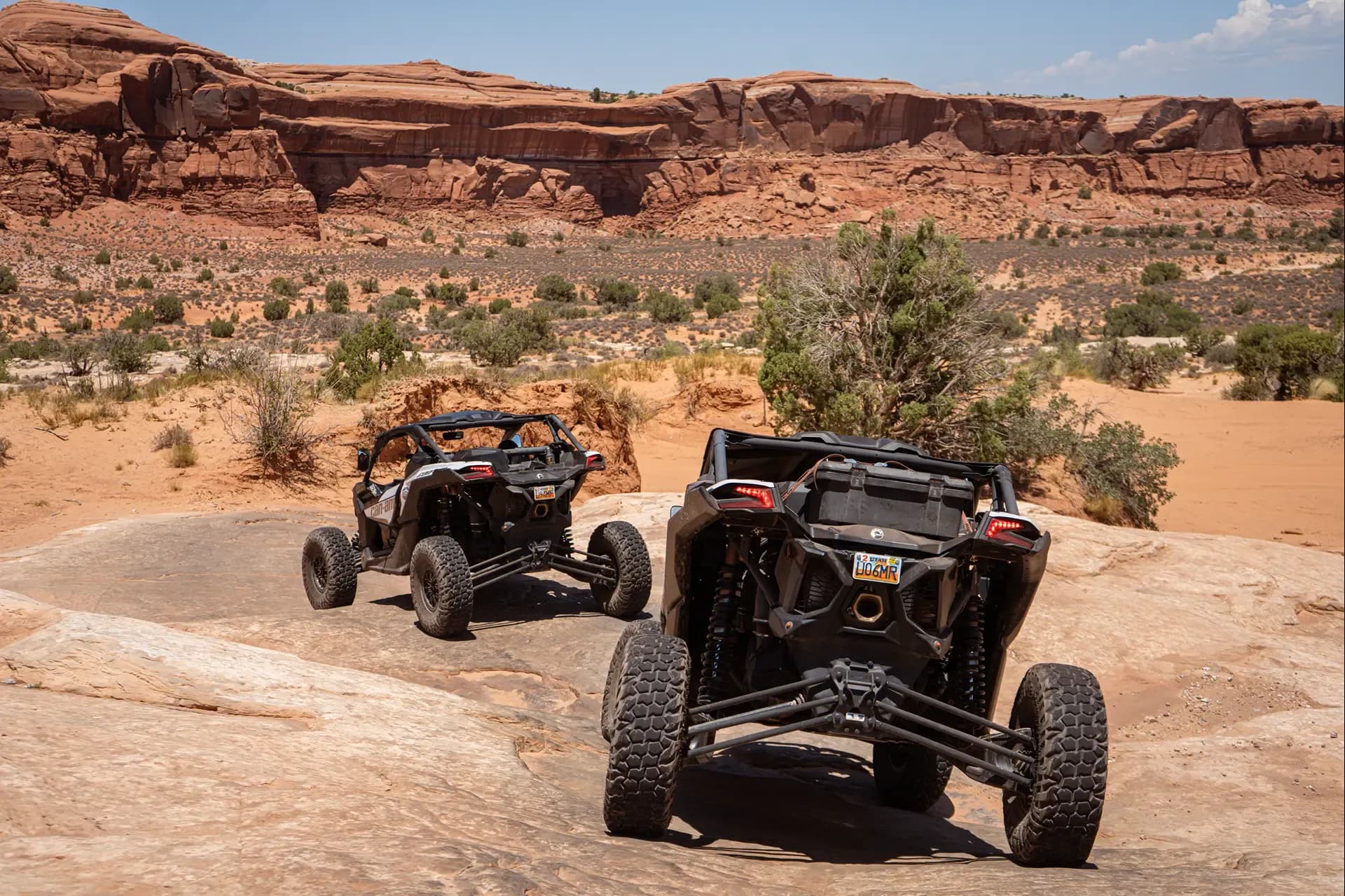 Two Can-Am side-by-sides parked on slickrock below a long red sandstone escarpment