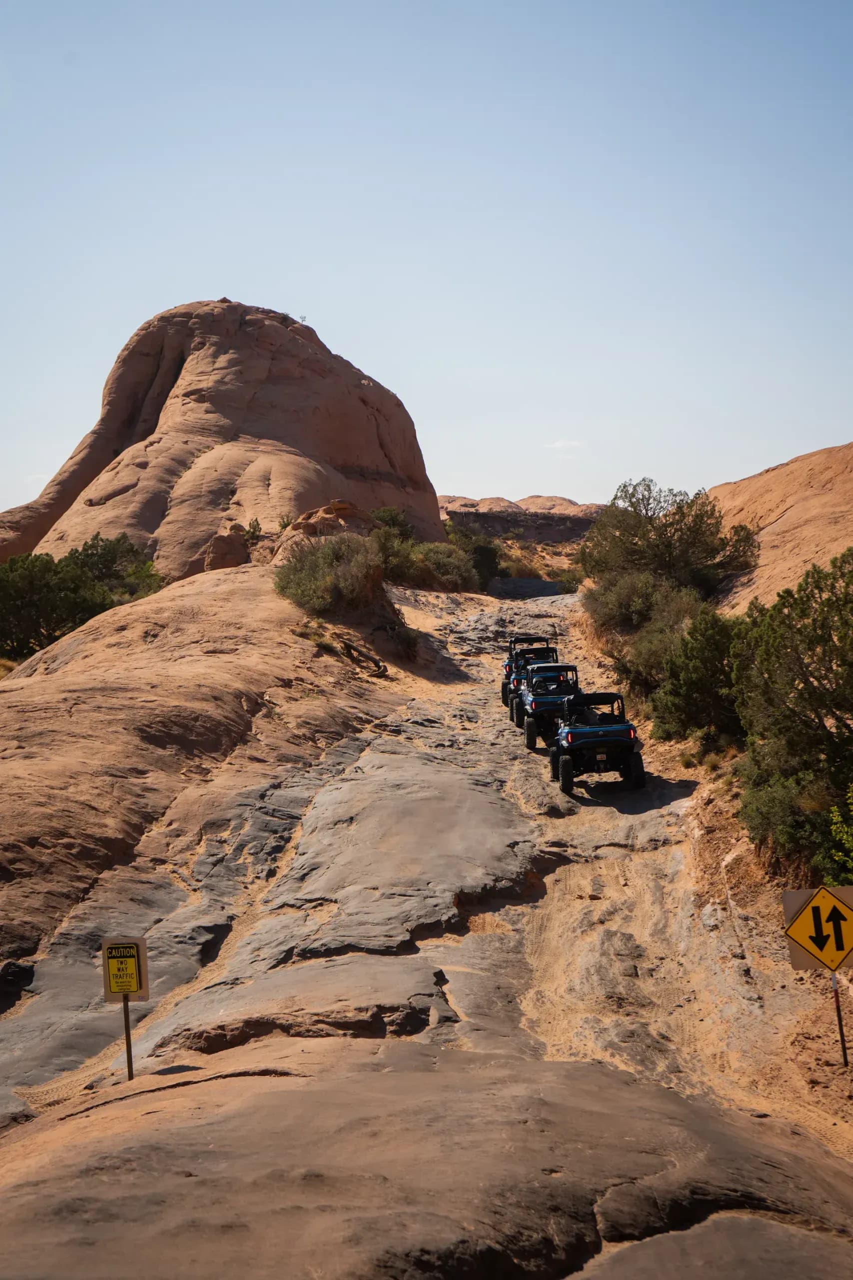 Convoy of UTVs climbing a rocky slickrock trail between sandstone walls and desert shrubs