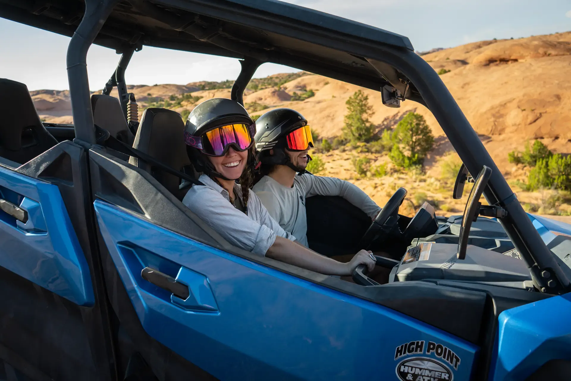 Smiling woman and man in helmets and goggles riding in a blue High Point side-by-side