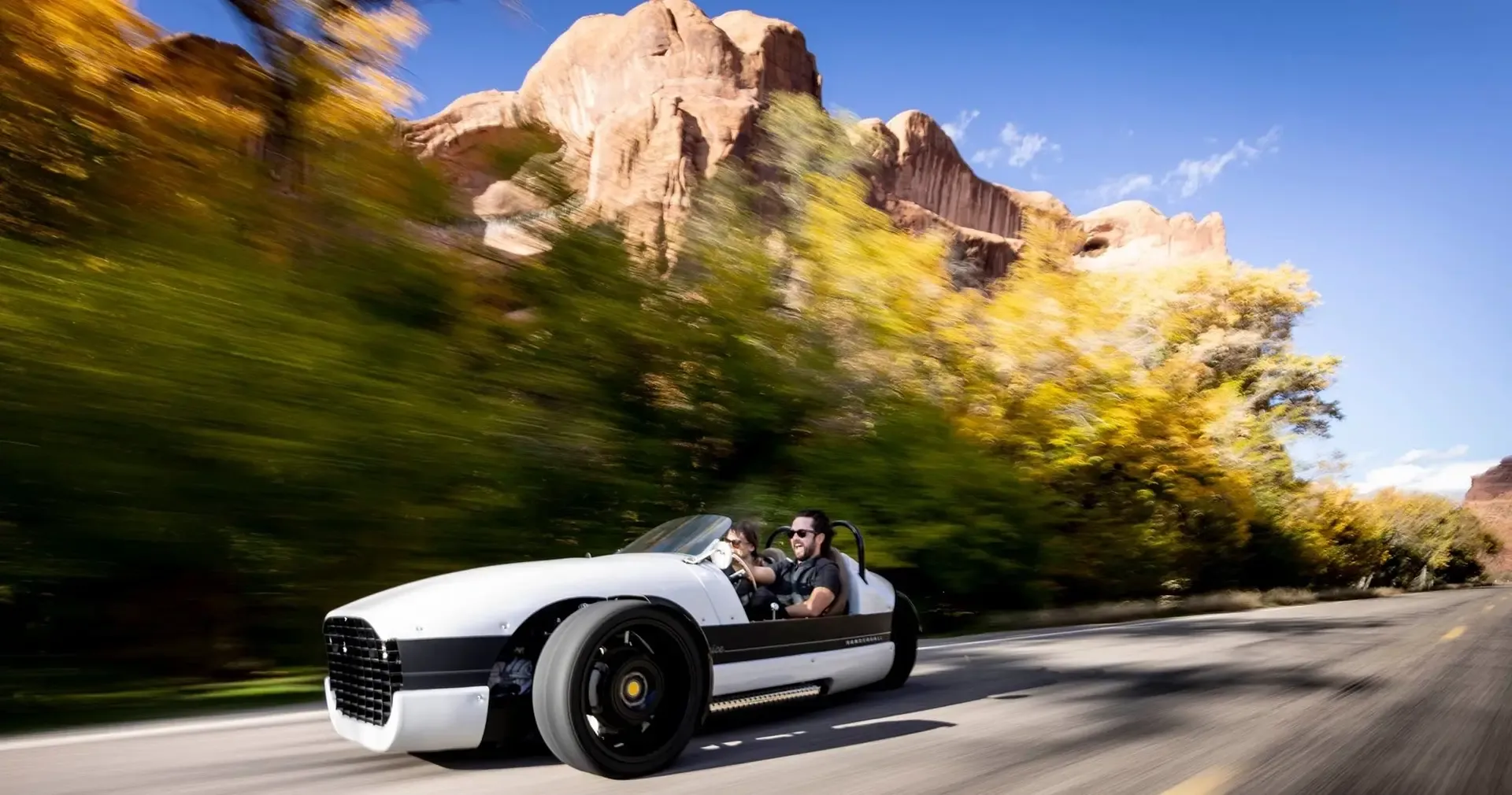 A white, three-wheeled roadster drives quickly down an asphalt road framed by yellow autumn trees and red rock cliffs.