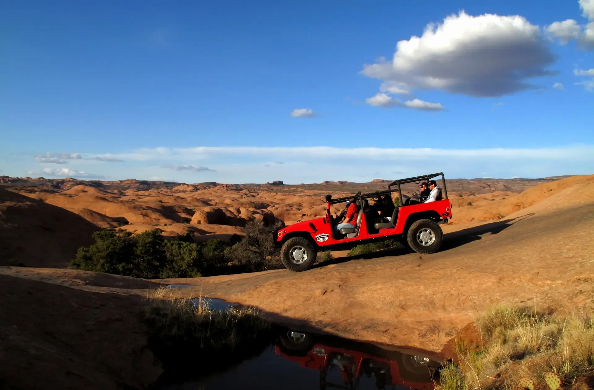 A red open-top hummer drives up a slickrock hill in a desert landscape under a blue sky with a single cloud.