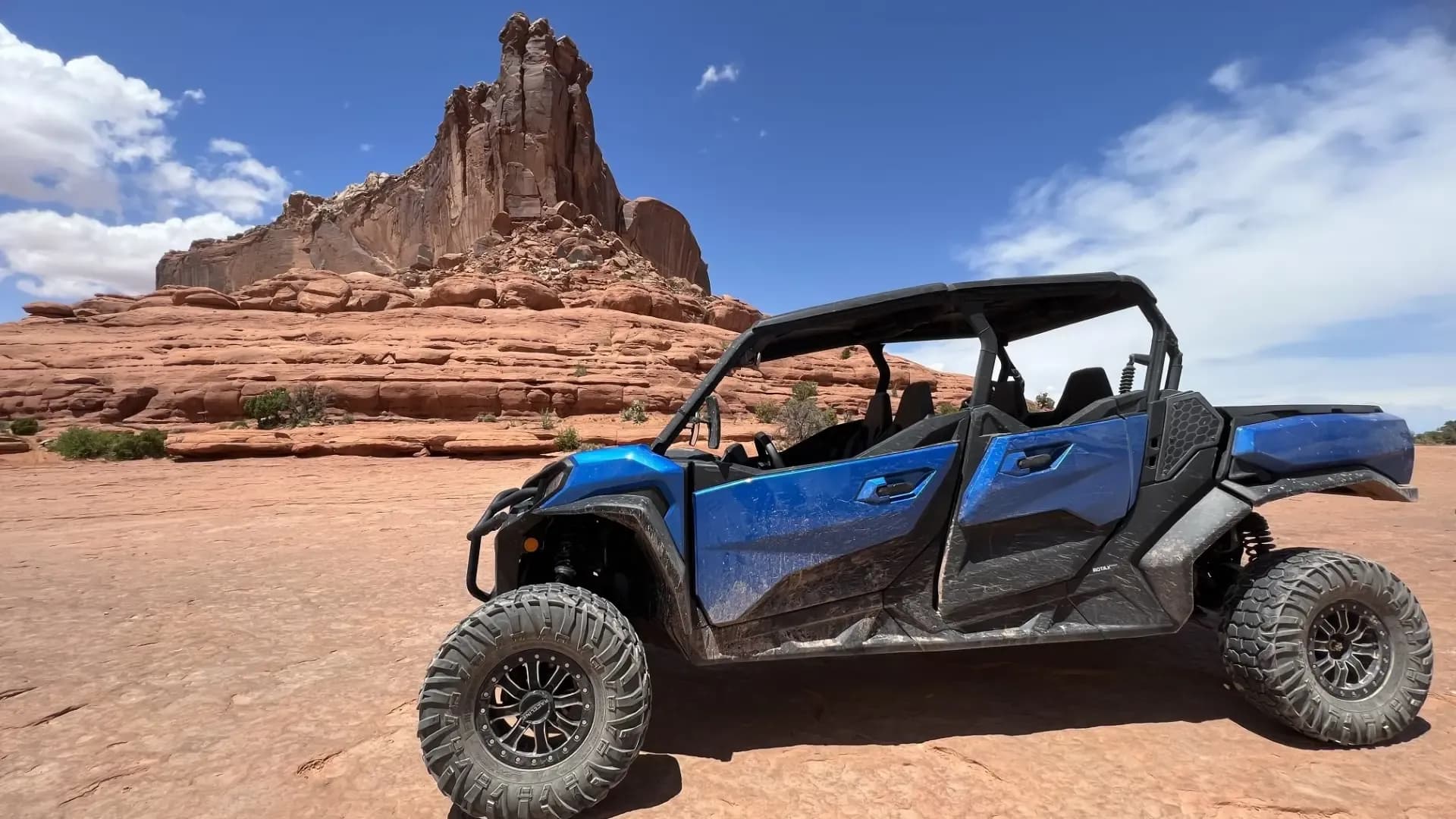 Blue four-seat side-by-side parked on red sand below a tall sandstone butte