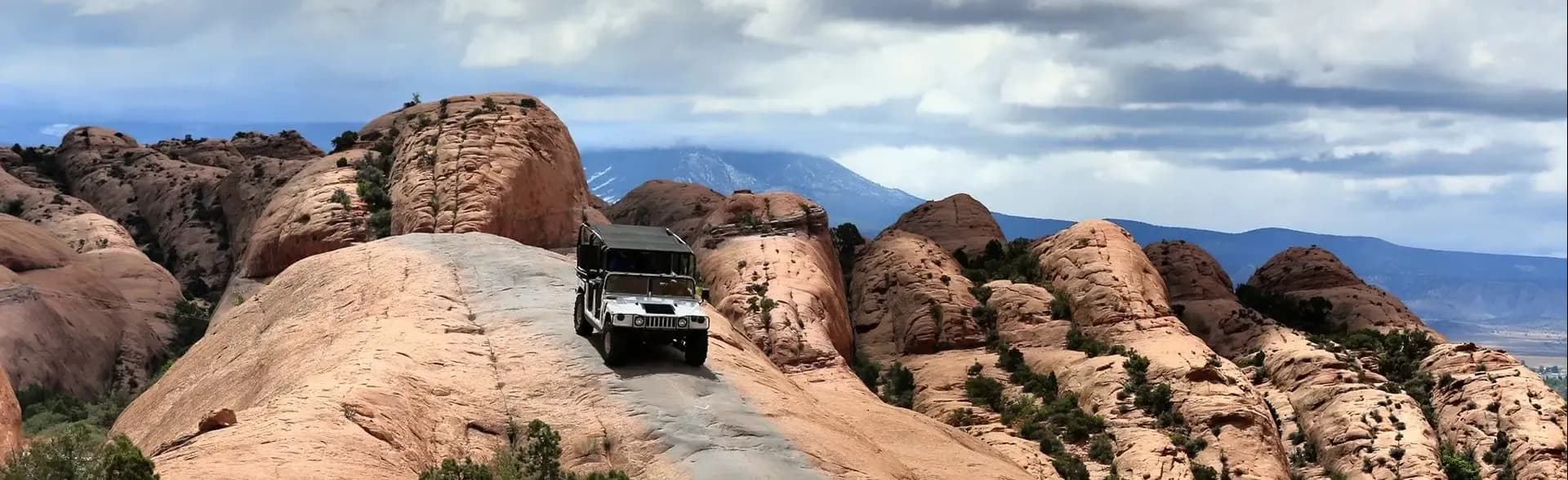 White Hummer on a slickrock ridge among sandstone fins with snowcapped mountains beyond