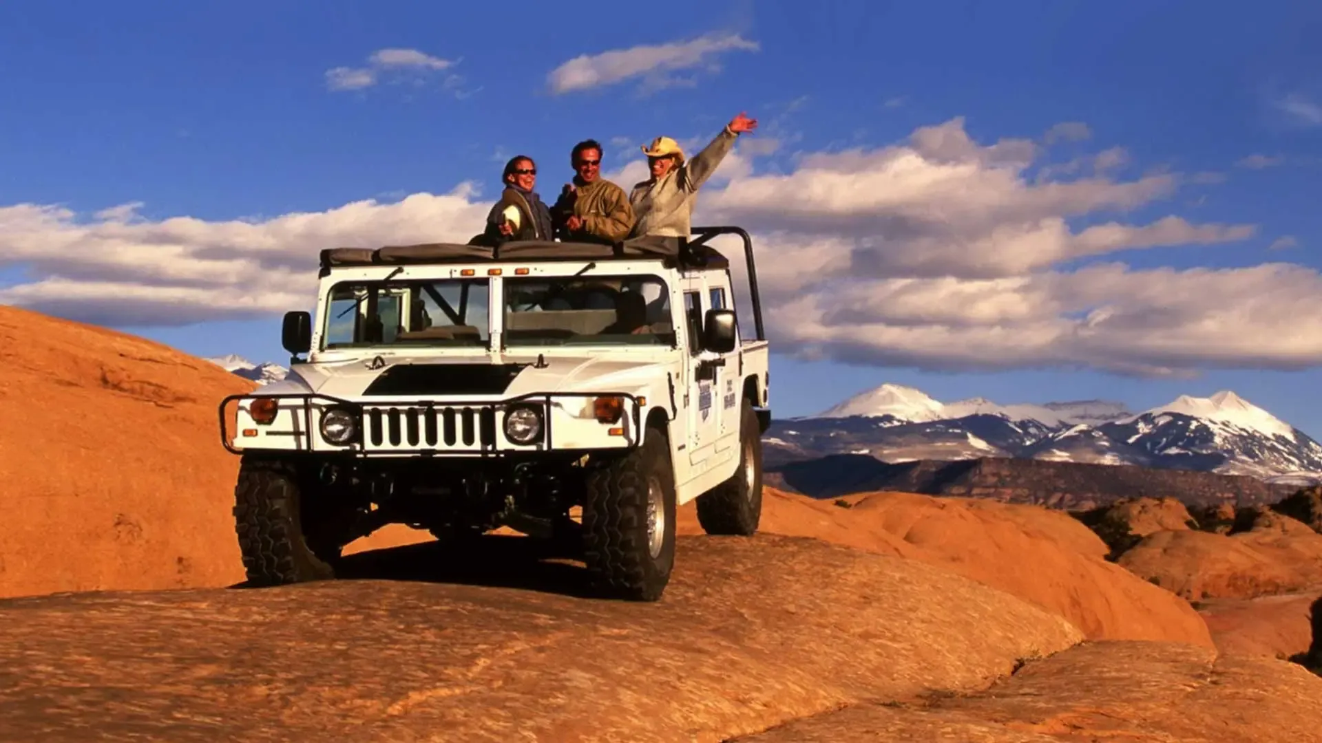Guests standing up and waving from a white Hummer on red rock with snowcapped mountains behind