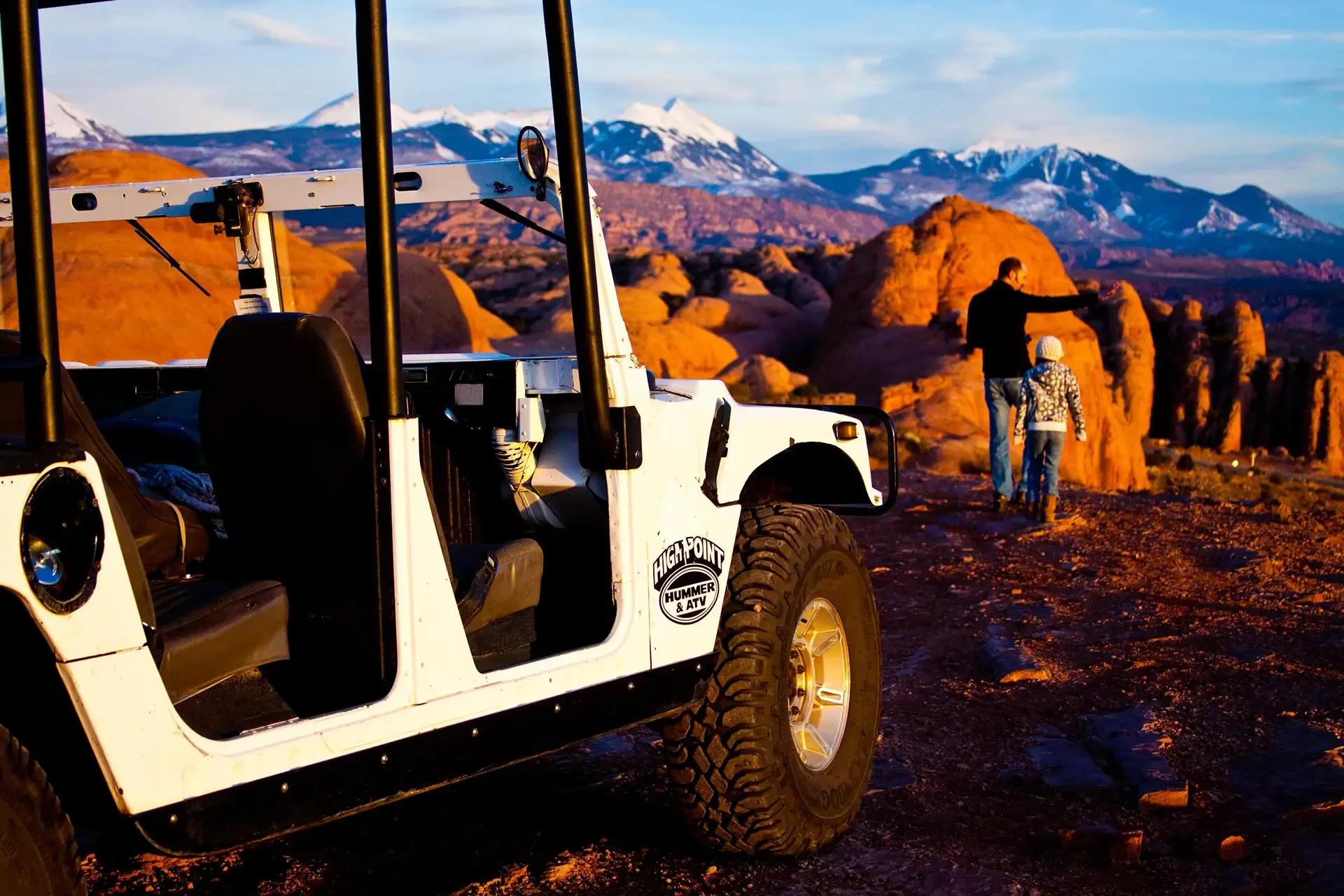 A white off-road vehicle is parked in the foreground as a man and child walk toward red rock formations at sunset.