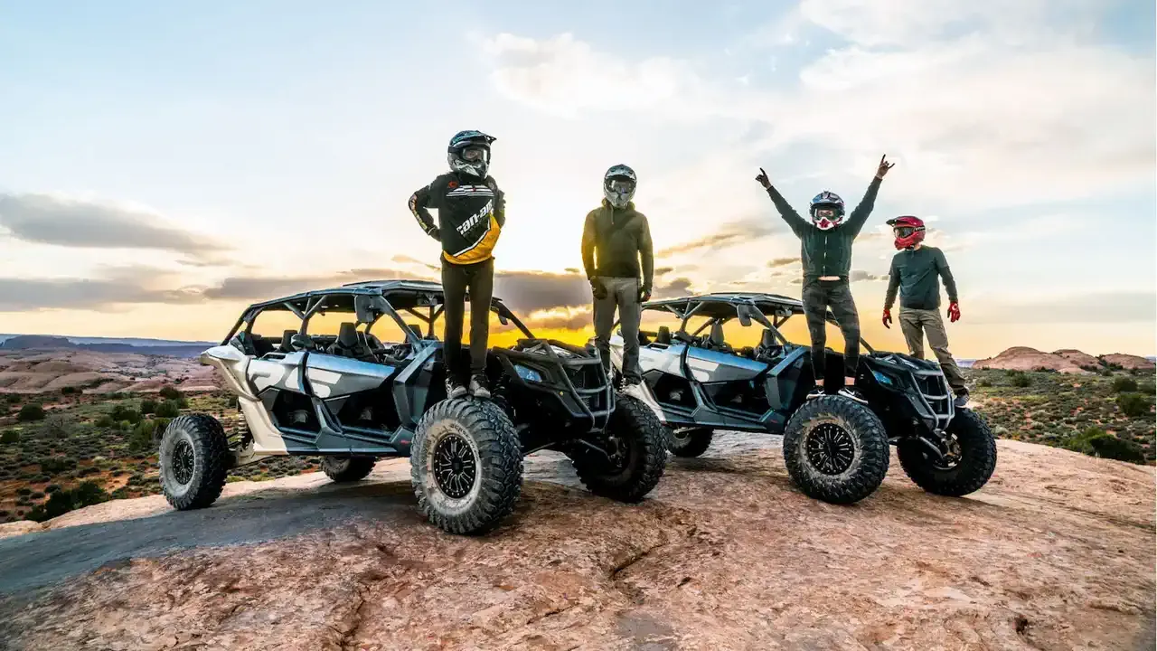 Four helmeted riders standing on top of two Can-Am side-by-sides on a rock outcrop, one with arms raised