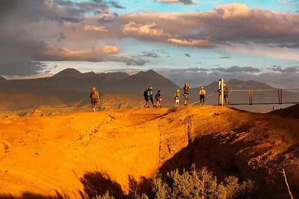 Group walking across a desert ridge at sunset toward the zipline launch platform
