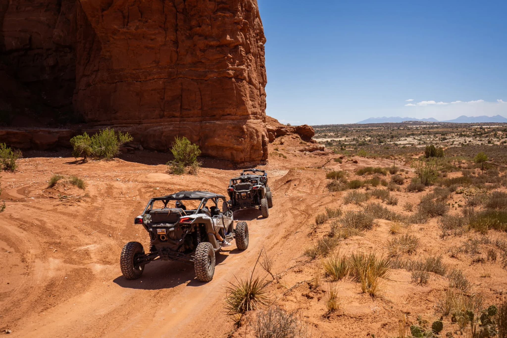 Two Can-Am side-by-sides driving a dirt trail at the foot of a towering red cliff