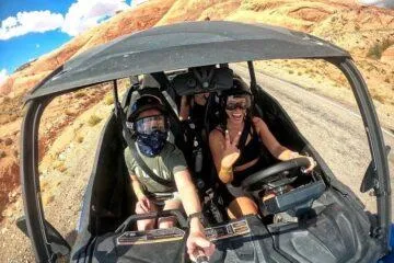 Two guests smiling for a selfie while driving a UTV on a desert trail