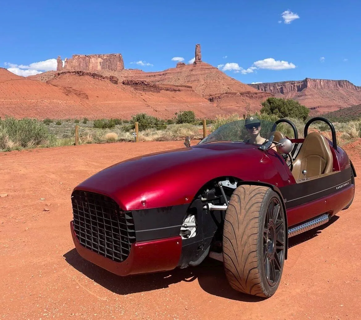 Red Vanderhall three-wheeled roadster parked on red dirt beneath towering sandstone buttes